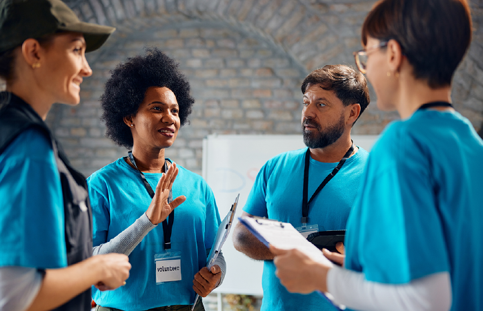Four people in matching blue volunteer shirts talk in a group, with some holding clipboards, in an indoor workspace.