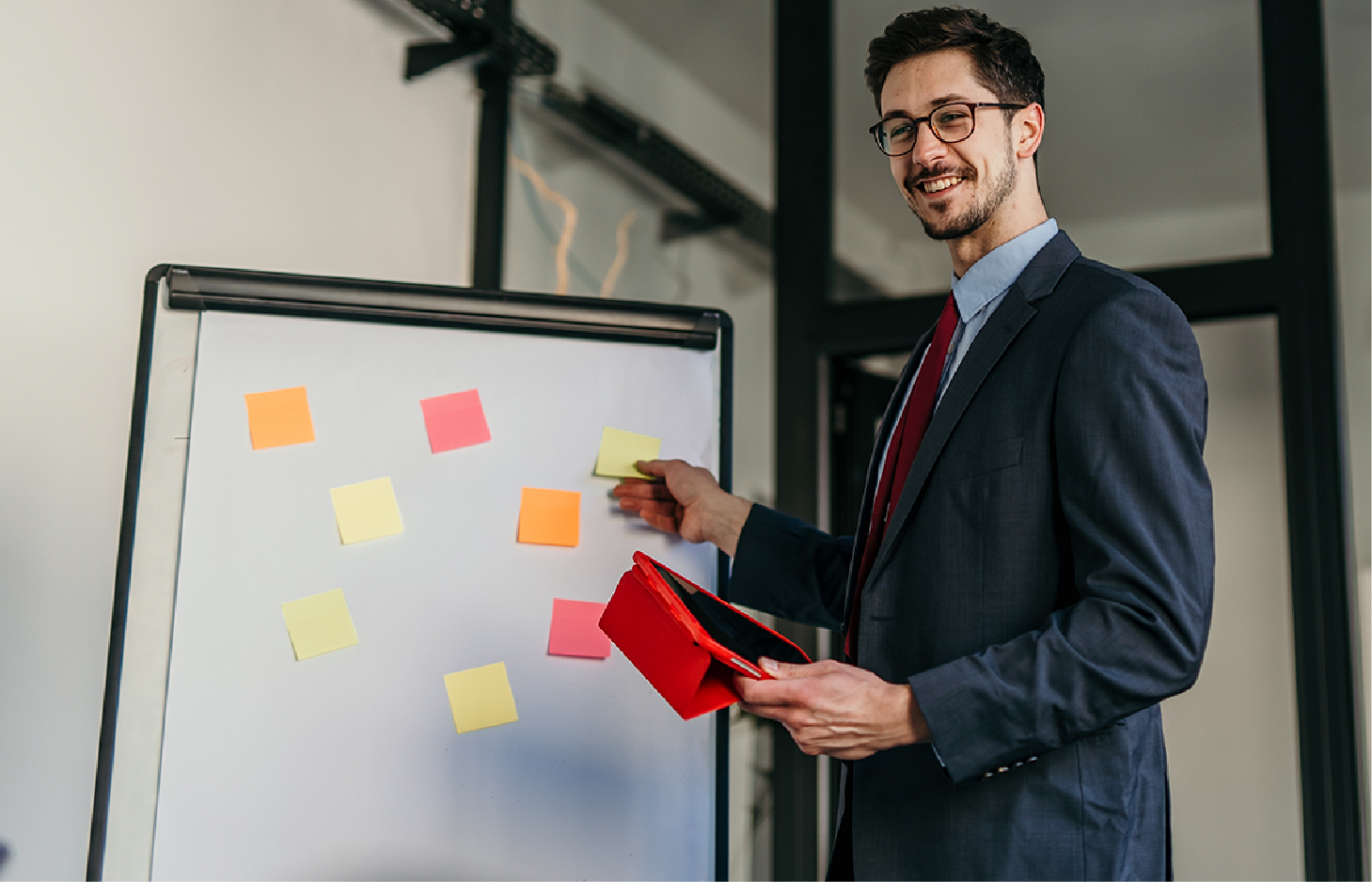 A professional in a suit smiles while pointing to colorful sticky notes on a whiteboard in an office.