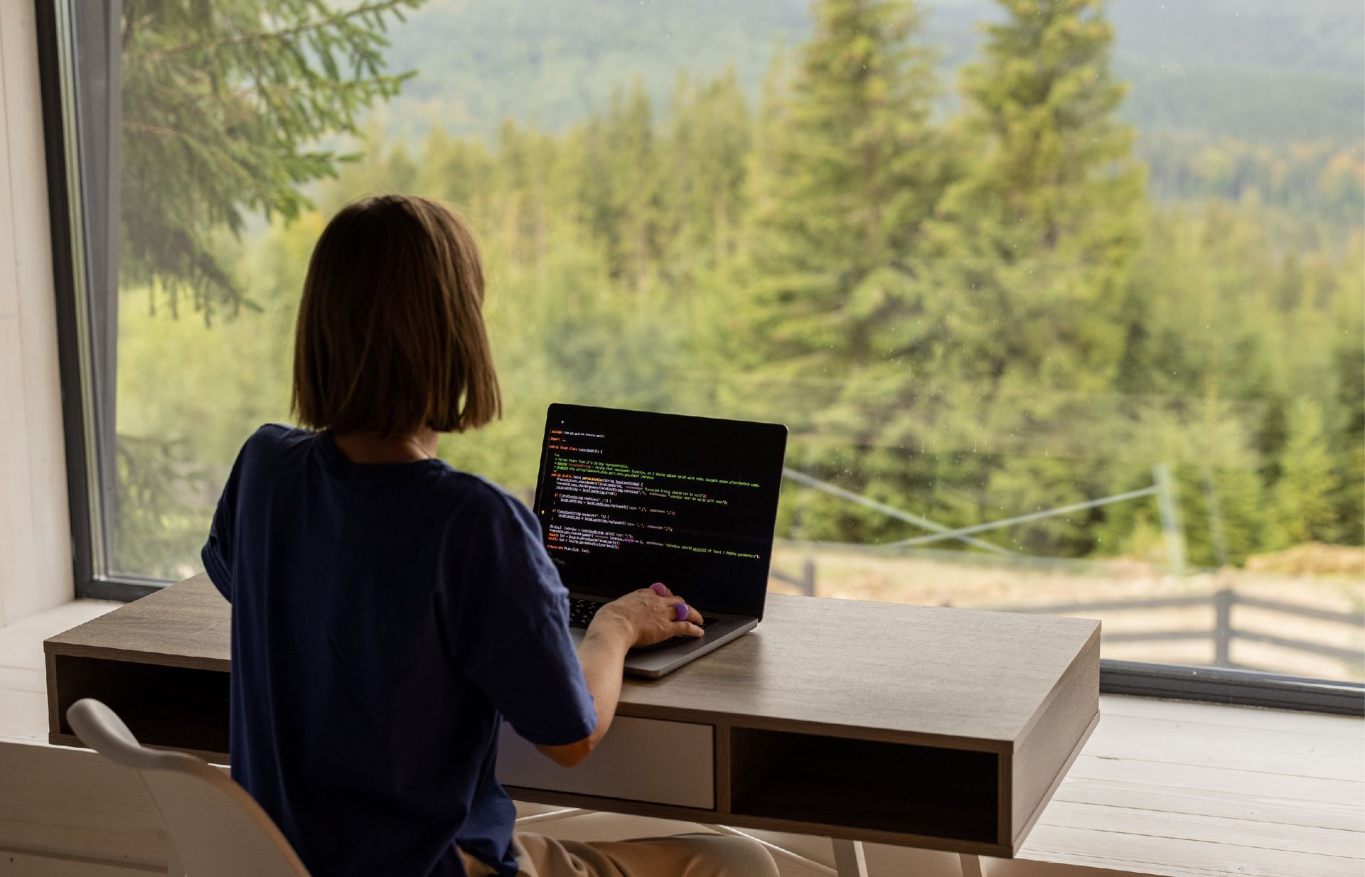 A person sits at a desk facing a window with a view of a forest, typing code on a laptop screen.