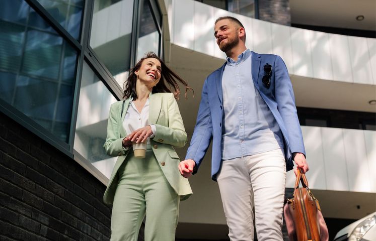 A smiling professional pair in business casual attire walking and talking outside a modern glass building.