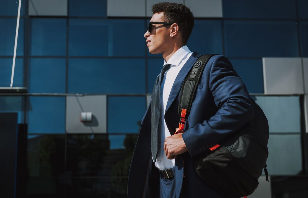 A person in a suit and sunglasses carries a black backpack while walking past a modern glass building.