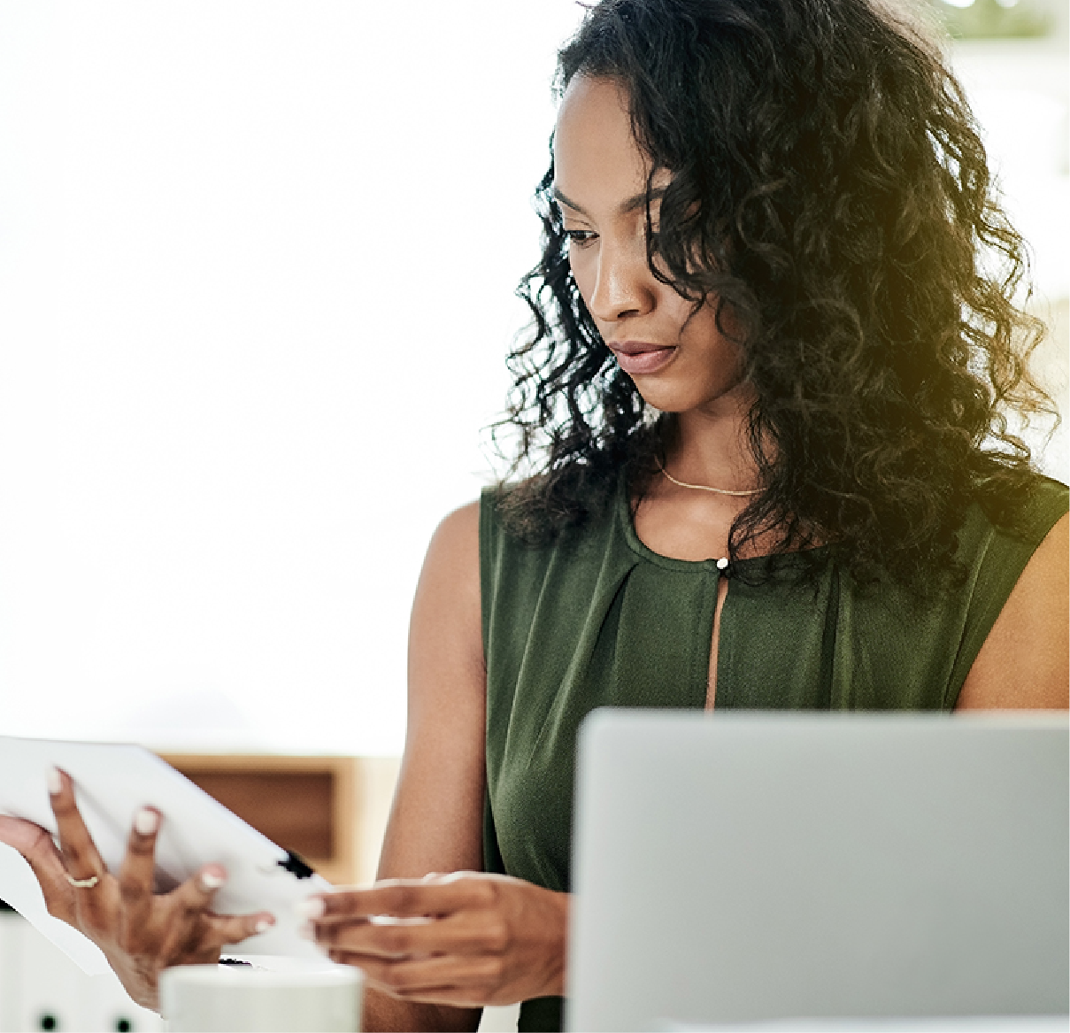 A focused professional in a green top holds papers while looking at a laptop in an office setting.