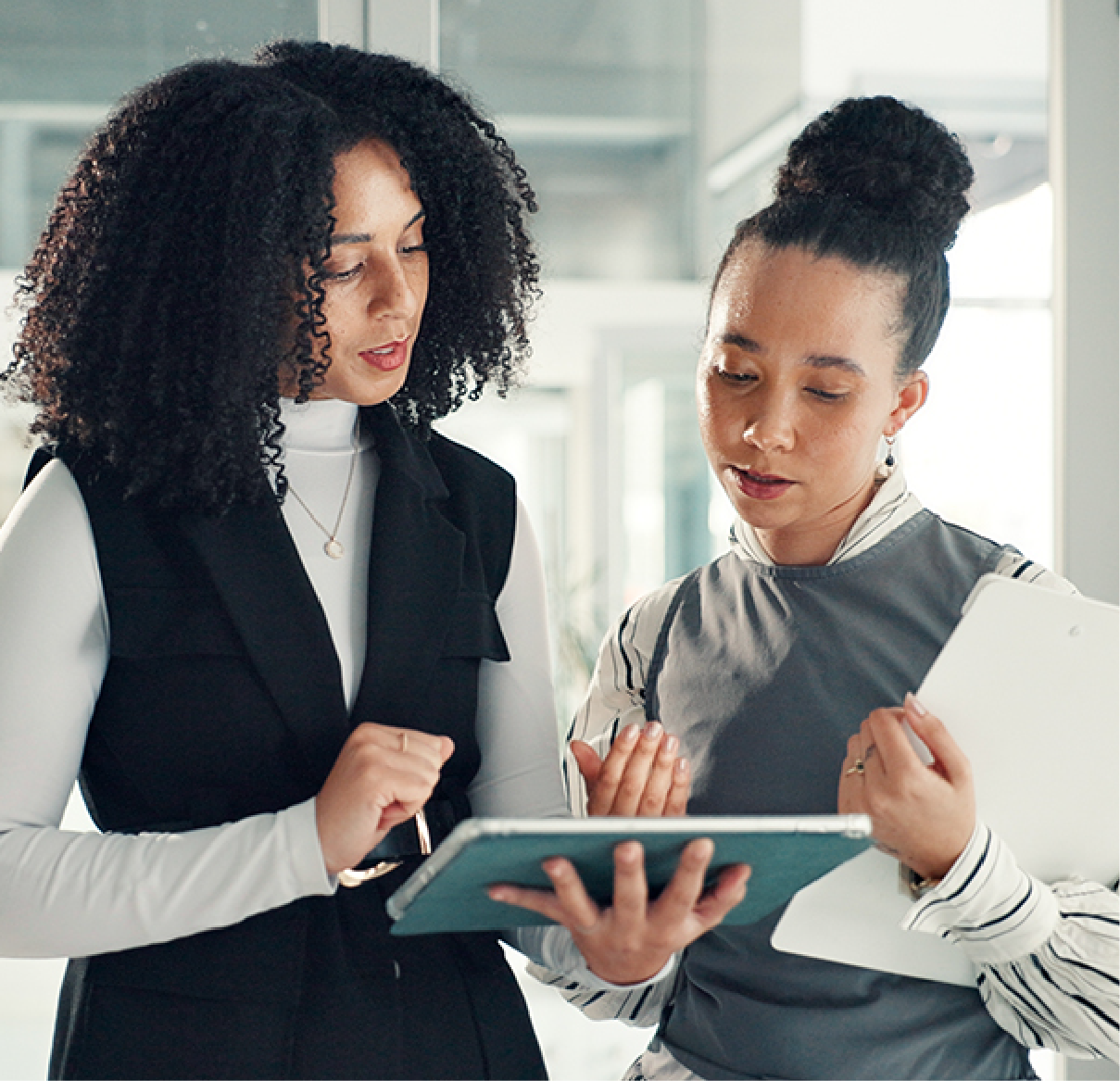 Two professionals stand together in an office, reviewing notes on a digital tablet and a paper file.