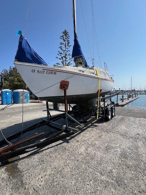 Sailboat on a trailer at a marina beside a dock, with blue covers and a clear sky.