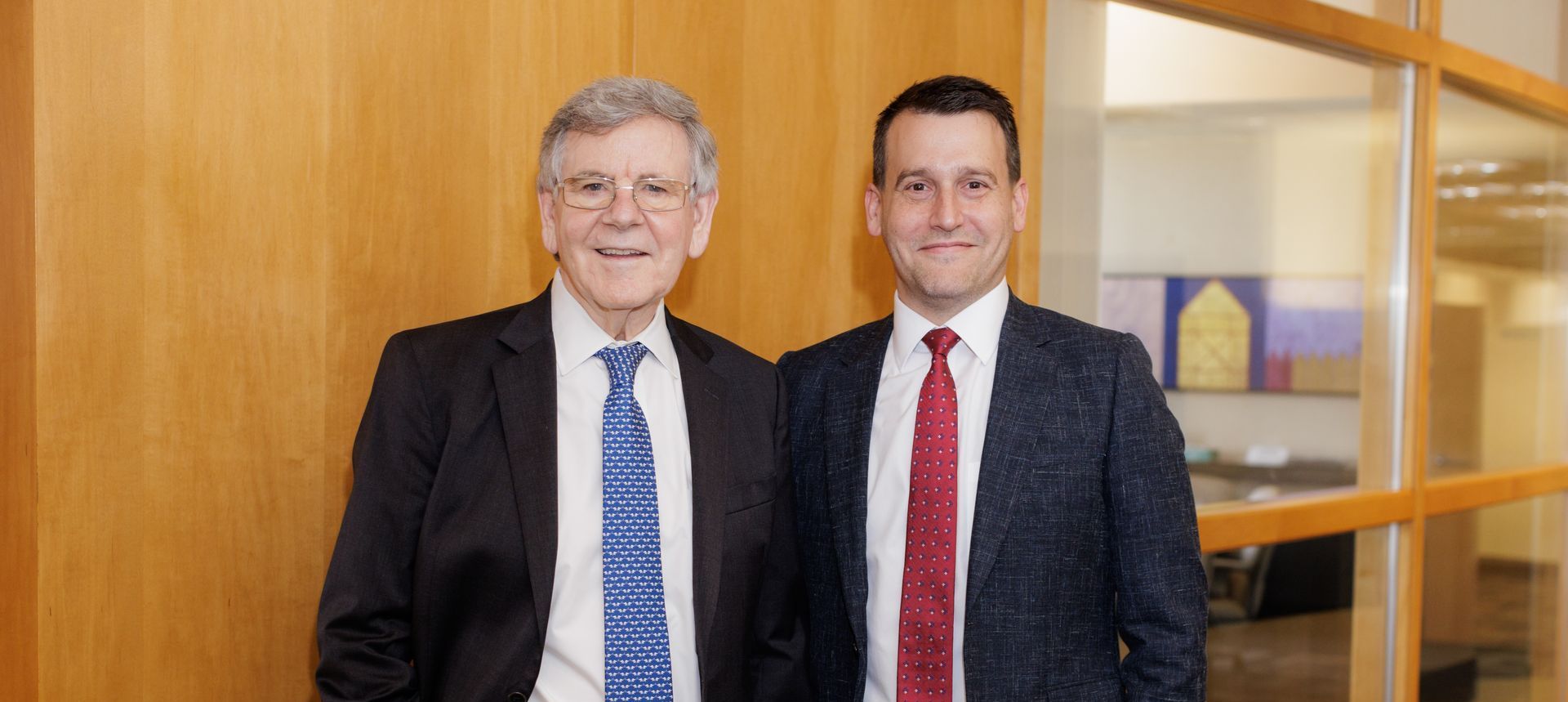 Two men in suits and ties are standing next to each other in front of a wooden wall.