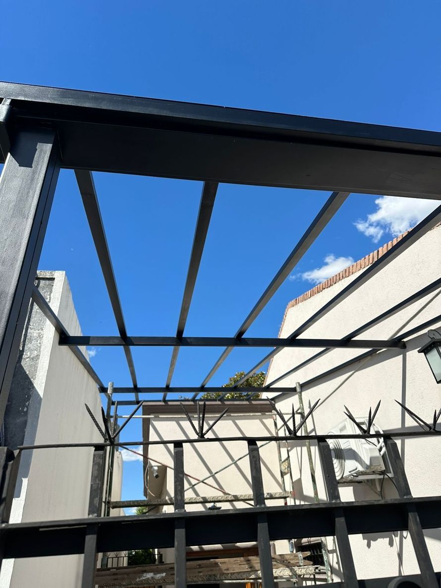 Patio abierto en la azotea con vigas de acero negro, paredes blancas y cielo azul.