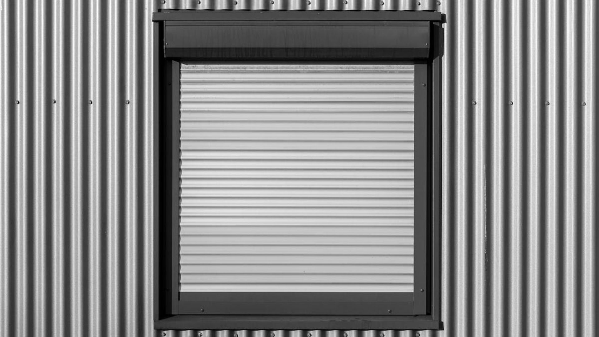 Persiana enrollable blanca cerrada, enmarcada en un marco oscuro sobre una pared de metal corrugado.