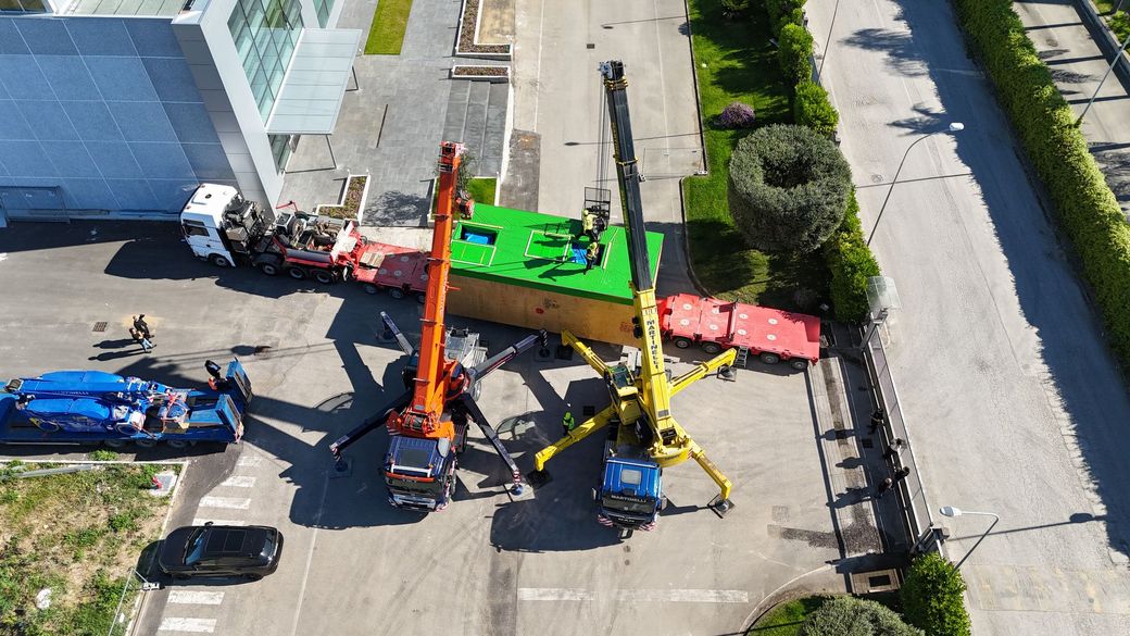 Veduta aerea di un cantiere edile con una gru, attrezzature colorate e operai vicino a un edificio e a una strada.