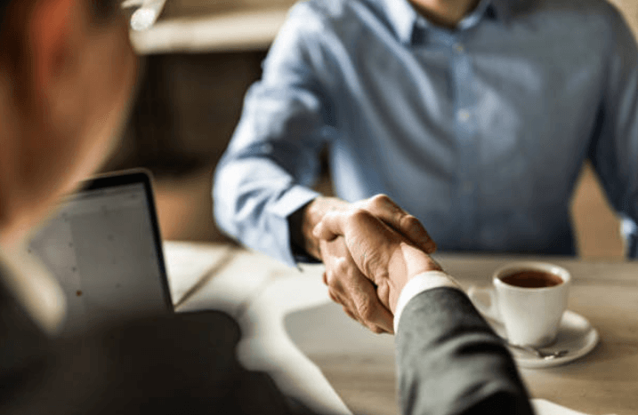 Two men are shaking hands over a table with a cup of coffee.