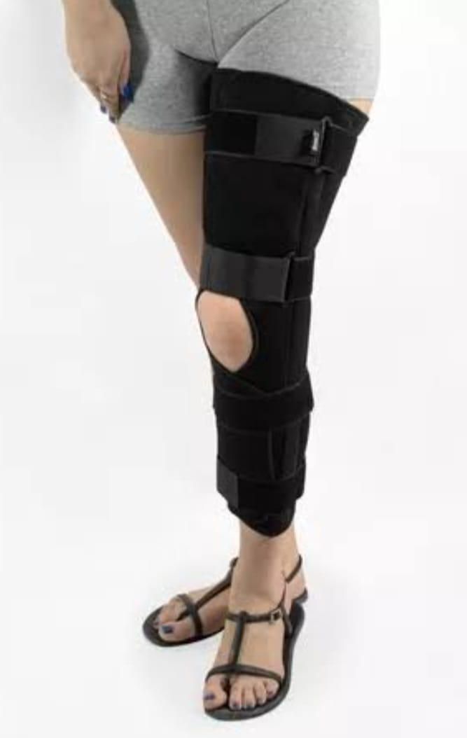 Persona que lleva una rodillera negra en una pierna, de pie con sandalias y pantalones cortos grises.