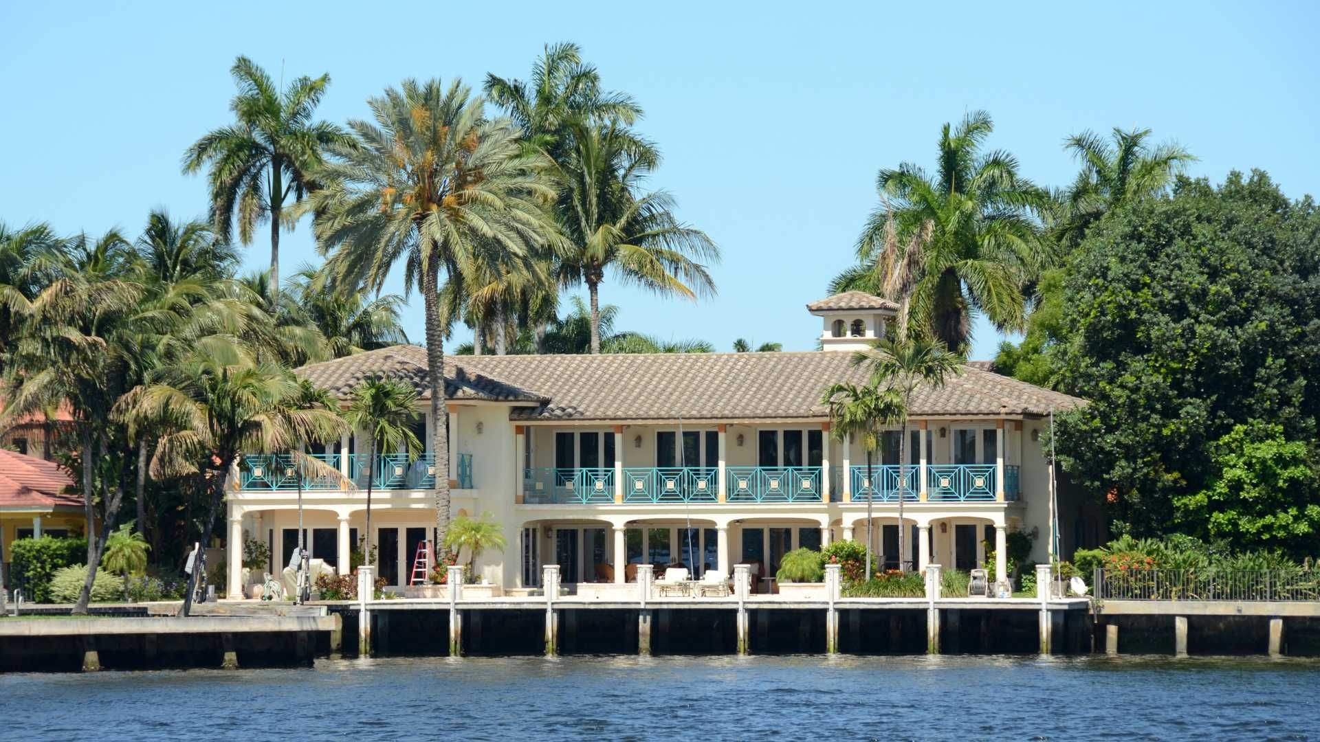 Waterfront mansion with beige facade, blue balconies, palm trees.