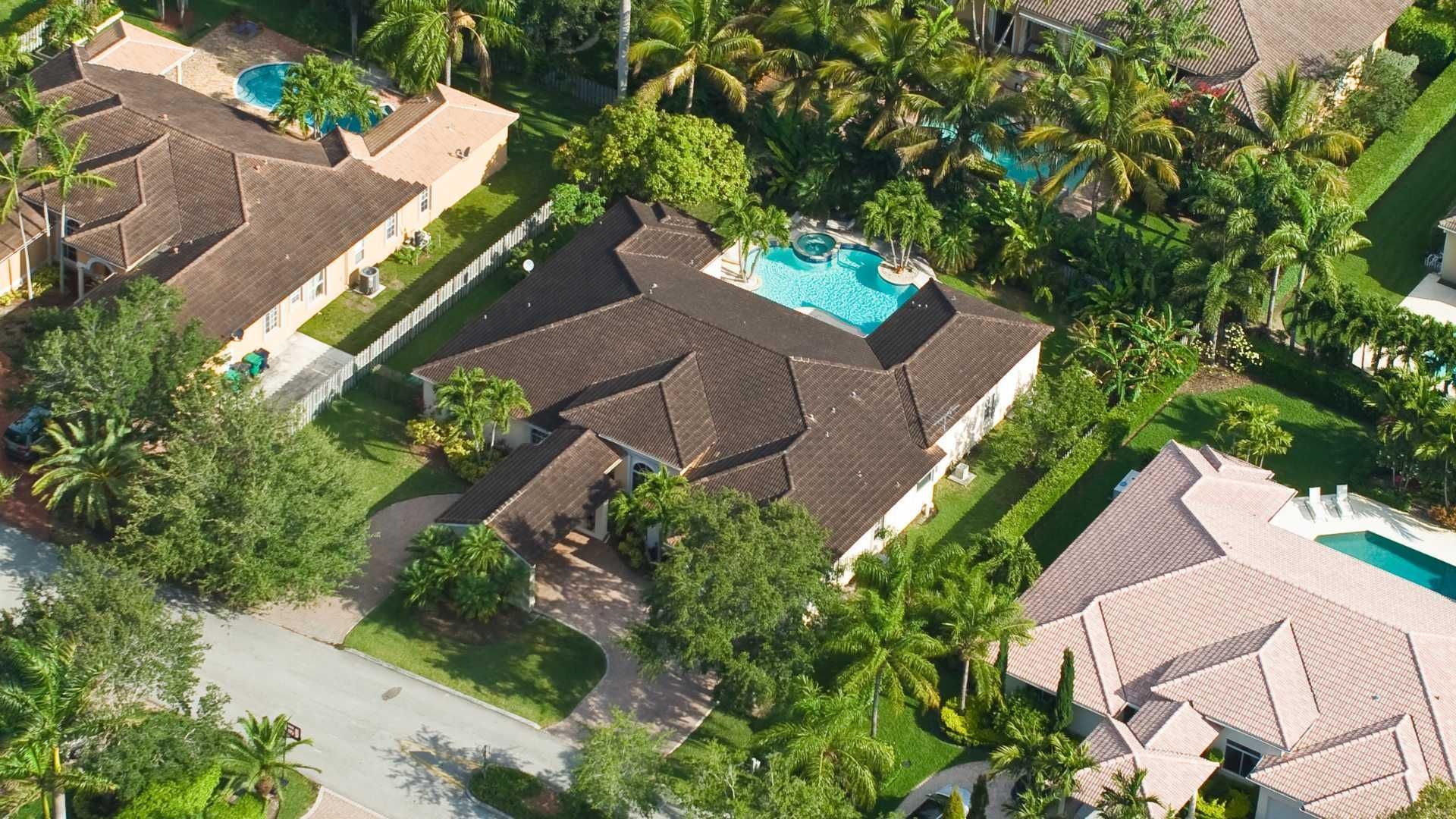 Aerial view of several houses with brown roofs, green trees, and blue swimming pools.