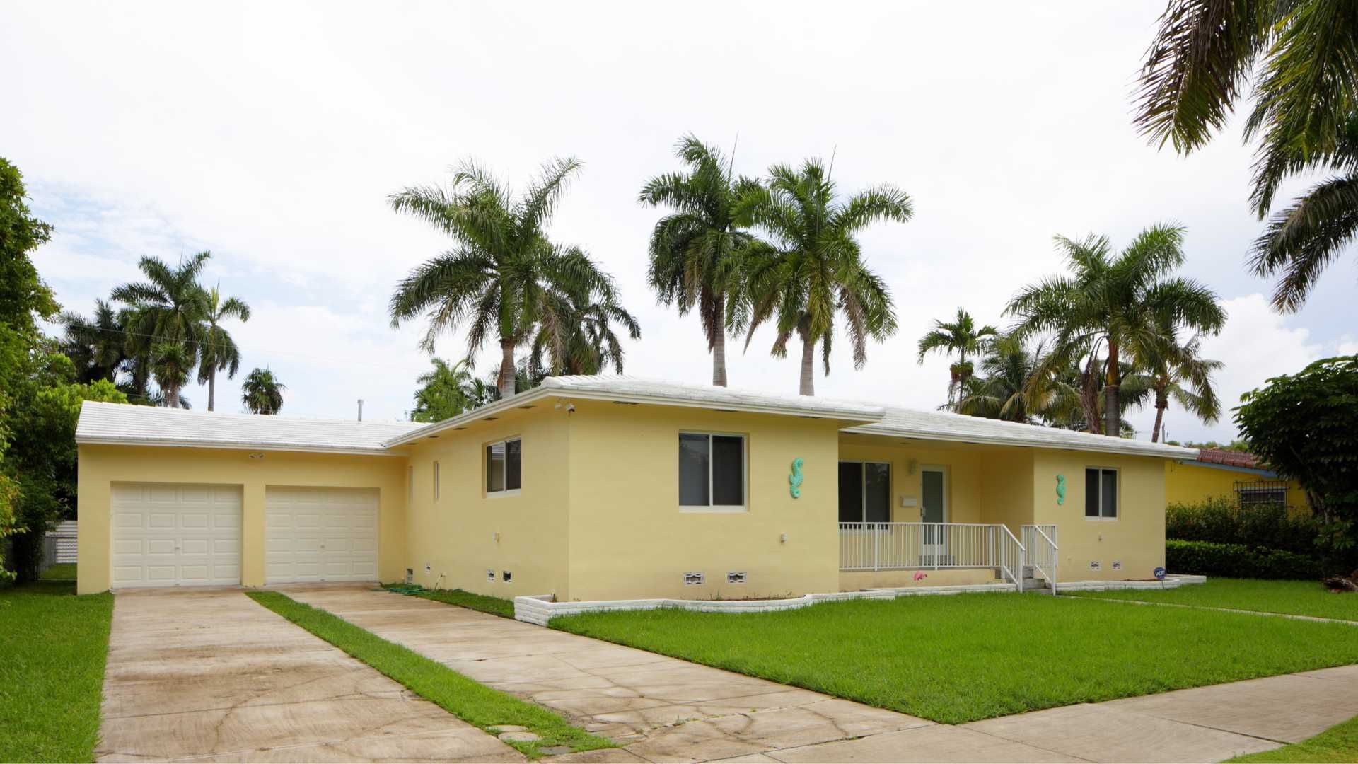 Yellow, single-story house with a two-car garage, green lawn, and palm trees in the background.