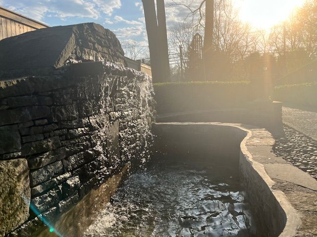 Stone waterfall cascading into a stone-lined pool. Bright sunlight filters through trees in the background.