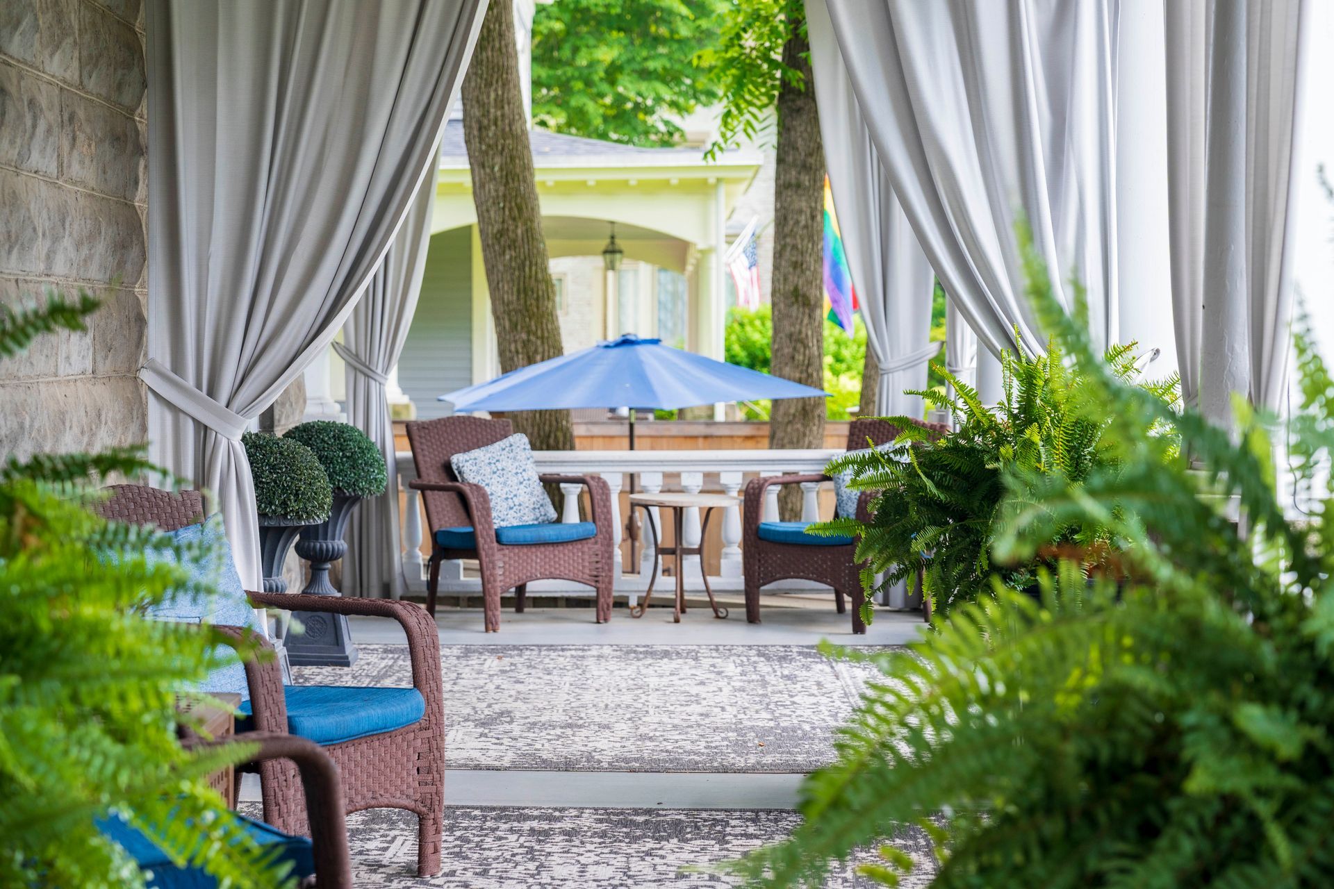 Covered porch with chairs, small table, and umbrella; ferns and curtains.