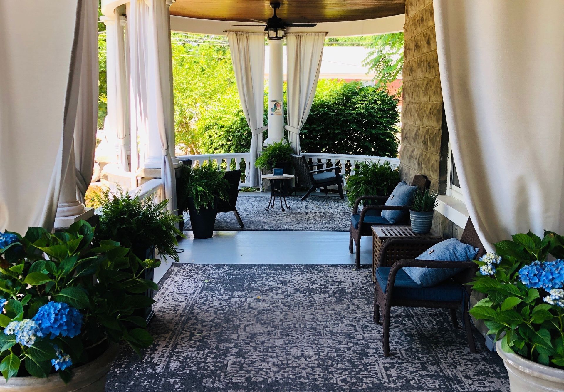 Covered porch with seating, plants, and white curtains; view of garden.