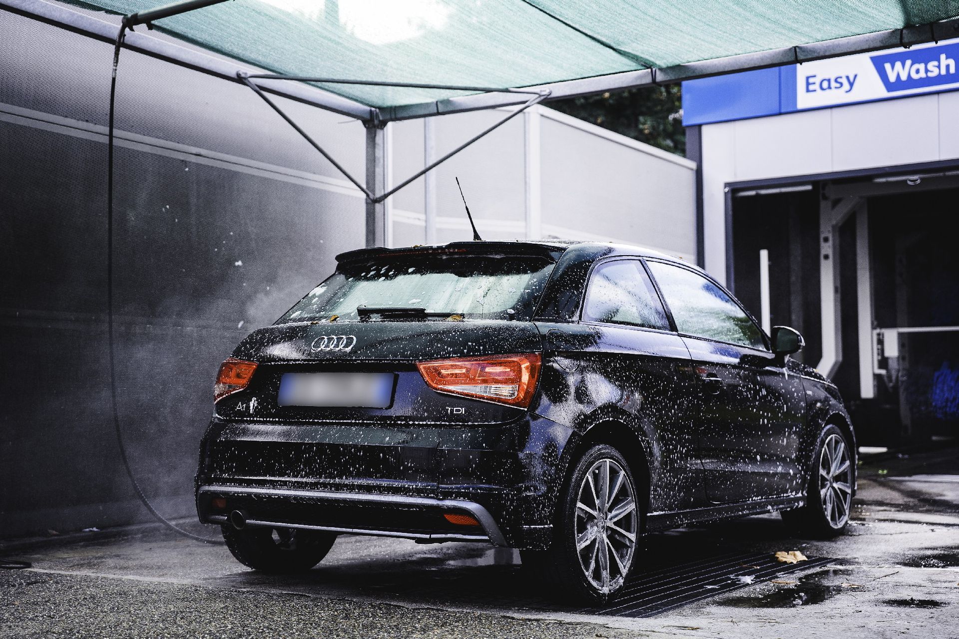 Un'Audi nera viene lavata in un autolavaggio Q8, con acqua e sapone visibili, sotto una tettoia.