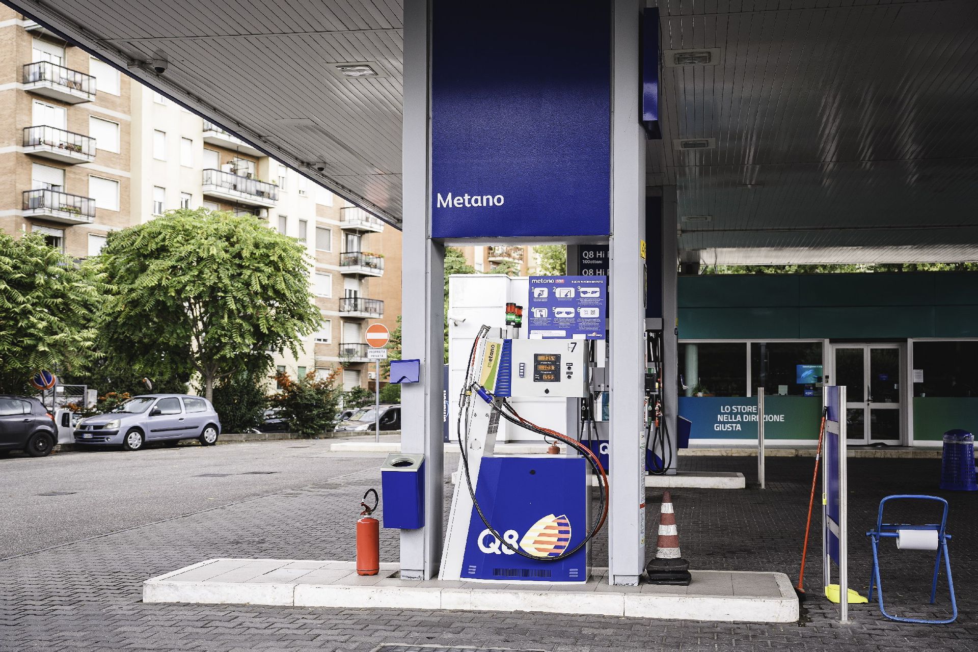 Stazione di servizio con pompa blu e bianca, sotto una tettoia. Auto e un edificio sullo sfondo.