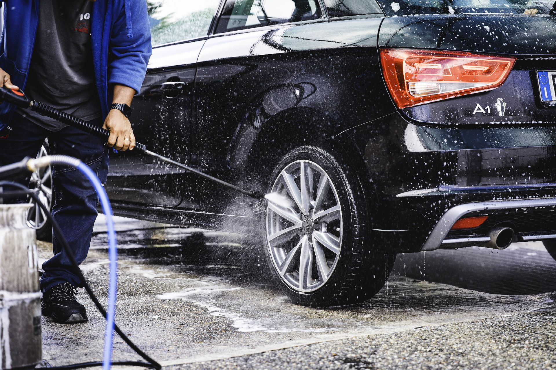Persona che lava un'auto nera con un'idropulitrice; spruzzi d'acqua visibili.