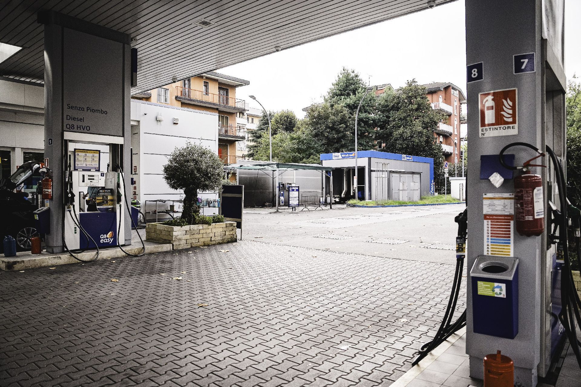 Esterno di una stazione di servizio con pompe di carburante, pavimentazione in mattoni ed edifici sullo sfondo.