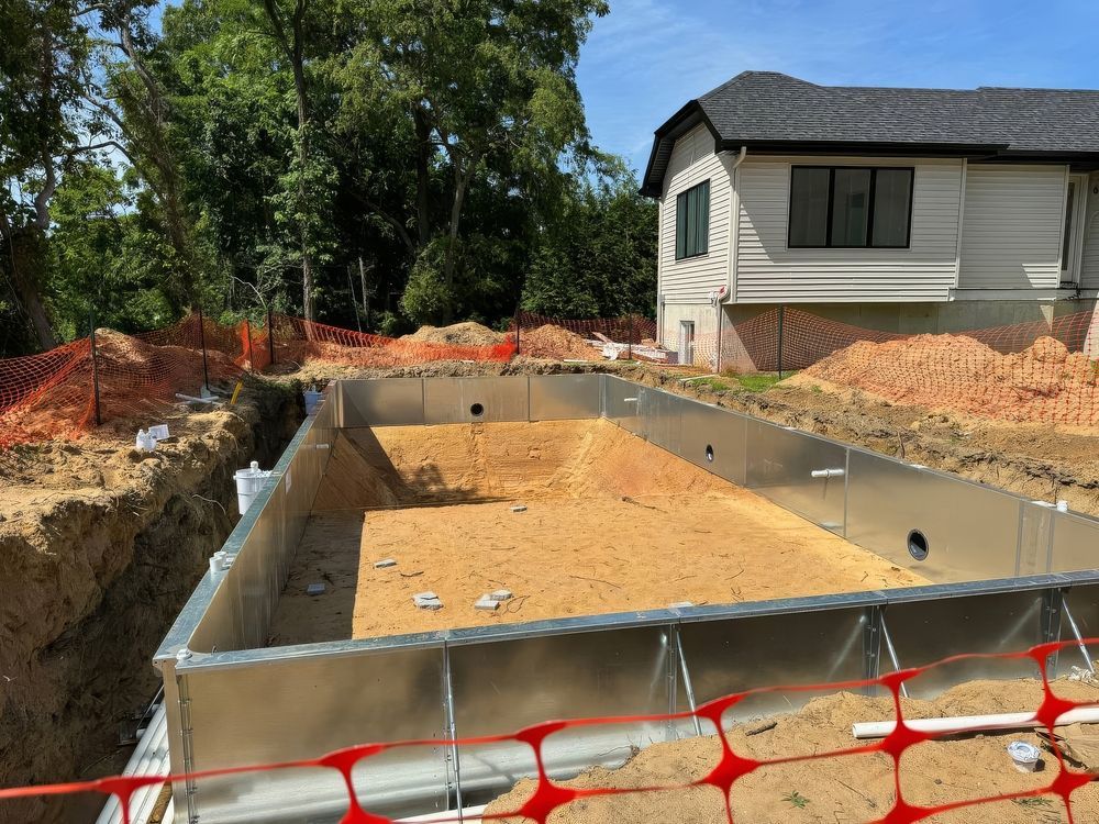 Concrete pool foundation under construction beside a white house, with orange safety fencing and dirt excavation.