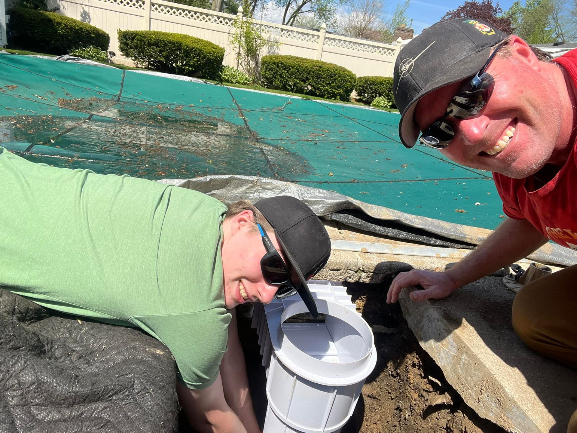 Two people crouch by a pool, peering into a white bucket in bright sunlight.