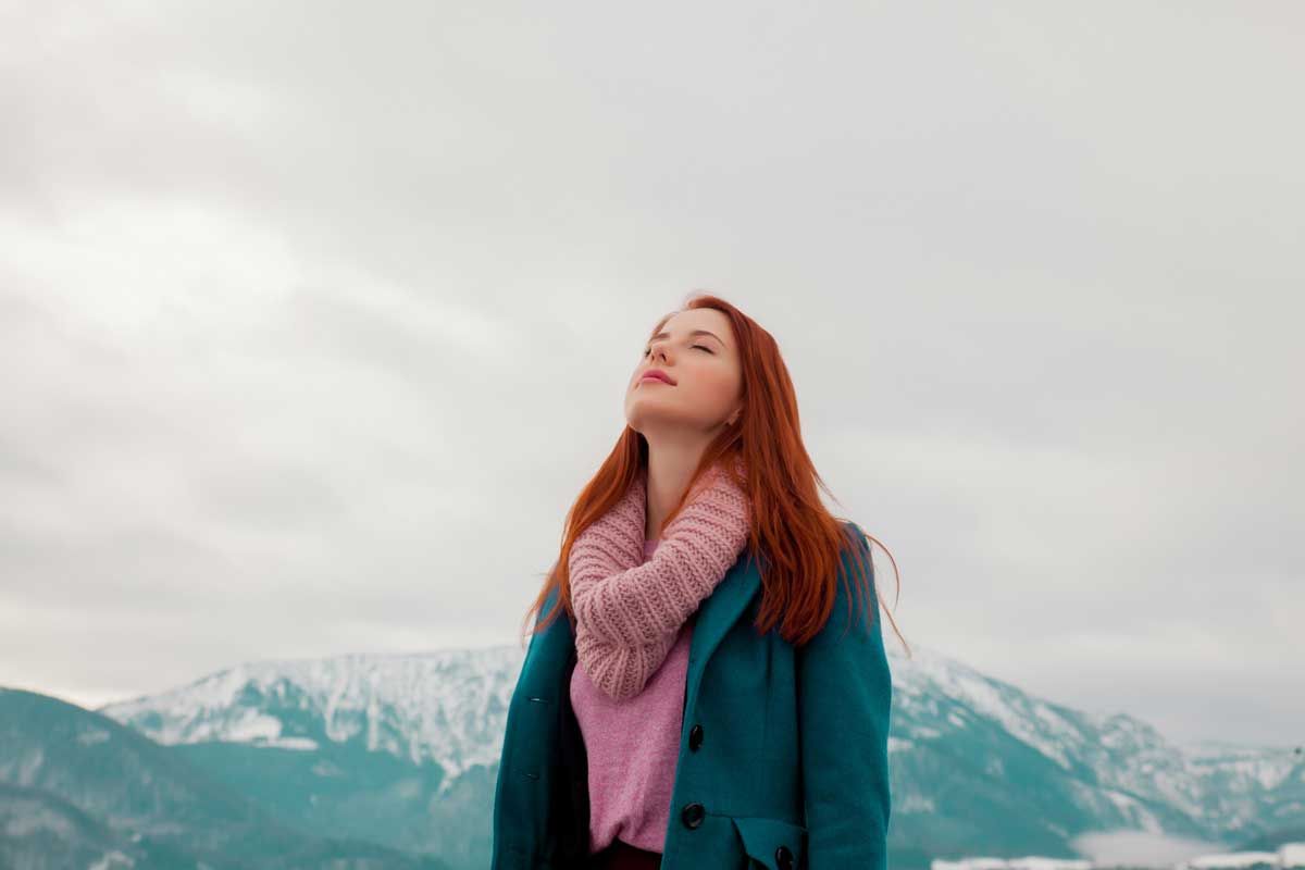 Woman with red hair, eyes closed, tilting head back, in a teal coat and pink scarf, with snowy mountains in background.