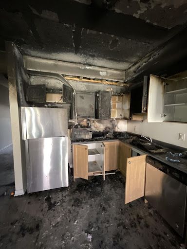 Fire-damaged kitchen: charred walls, appliances, cabinets. Debris on floor.