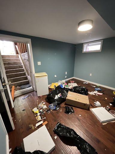 Messy basement room with dark wood floor, garbage bags, trash, small fridge, and stairs. Teal walls.
