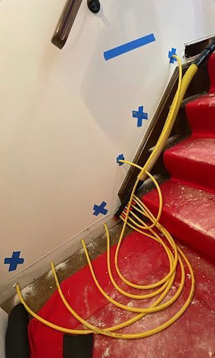 Yellow electrical cord coiled on red-carpeted stairs with white walls, blue tape markers, and brown banister.