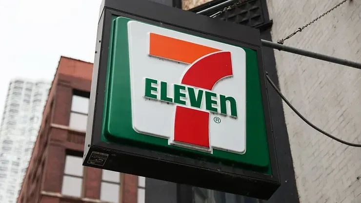 A 7 eleven sign is hanging from the side of a building.