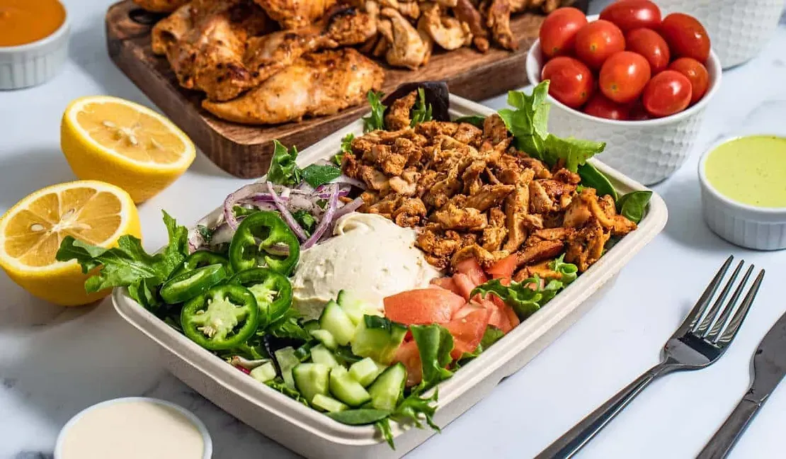 A container of food with chicken , tomatoes , cucumbers , onions and lemons on a table.