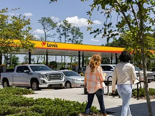 Two women are walking in front of a gas station.