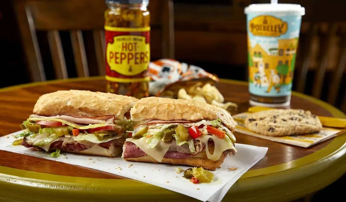 A sub sandwich is sitting on a table next to a bottle of hot pepper.