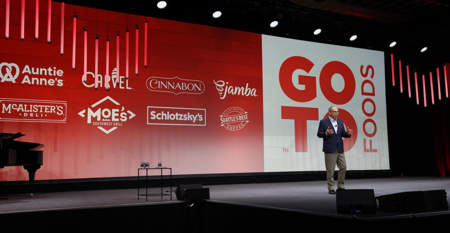 A man is standing on a stage in front of a large screen that says go to foods.