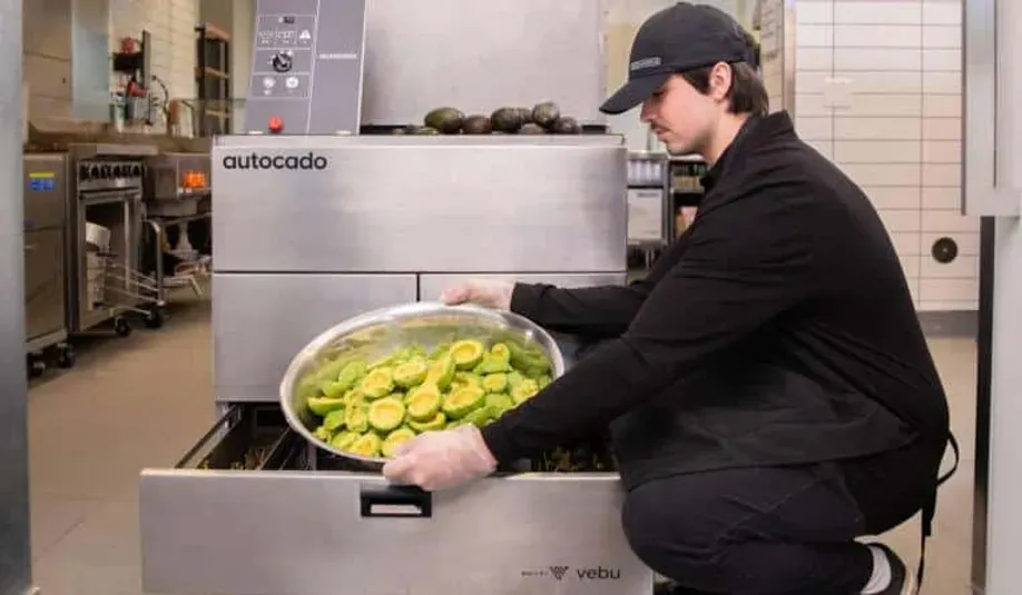 A man is kneeling down in a kitchen holding a bowl of avocados.
