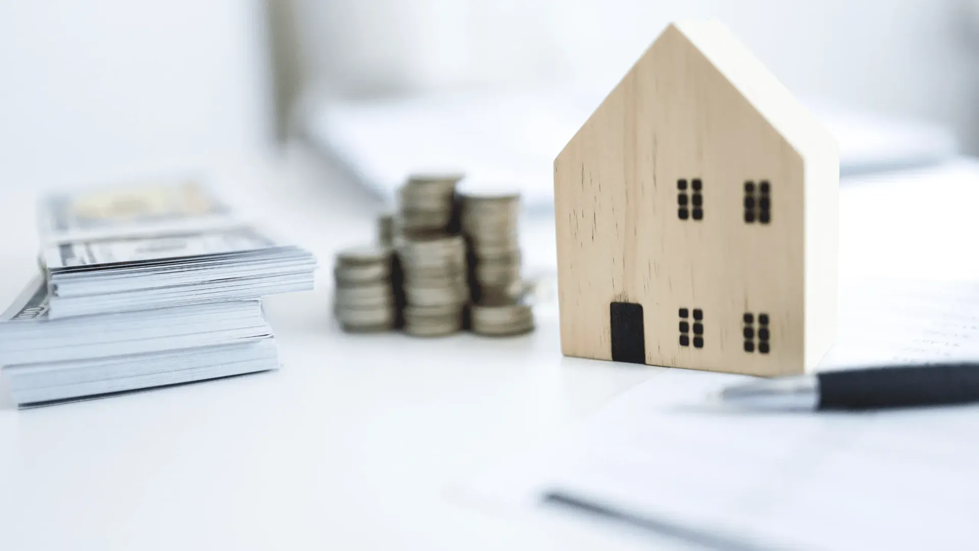 A small wooden house is sitting next to a pile of coins on a table.
