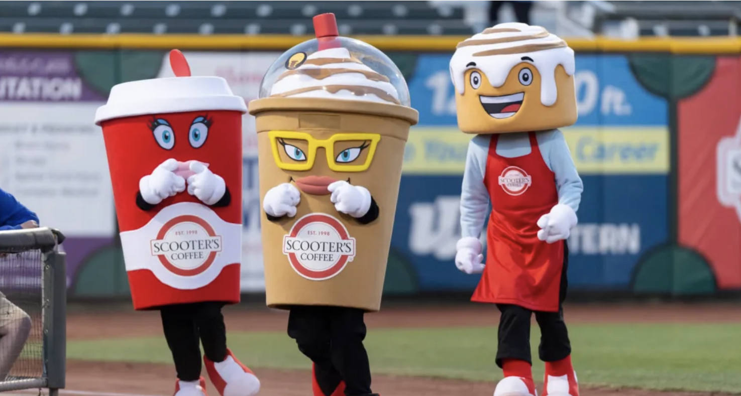 Three mascots dressed as coffee cups are standing on a baseball field.