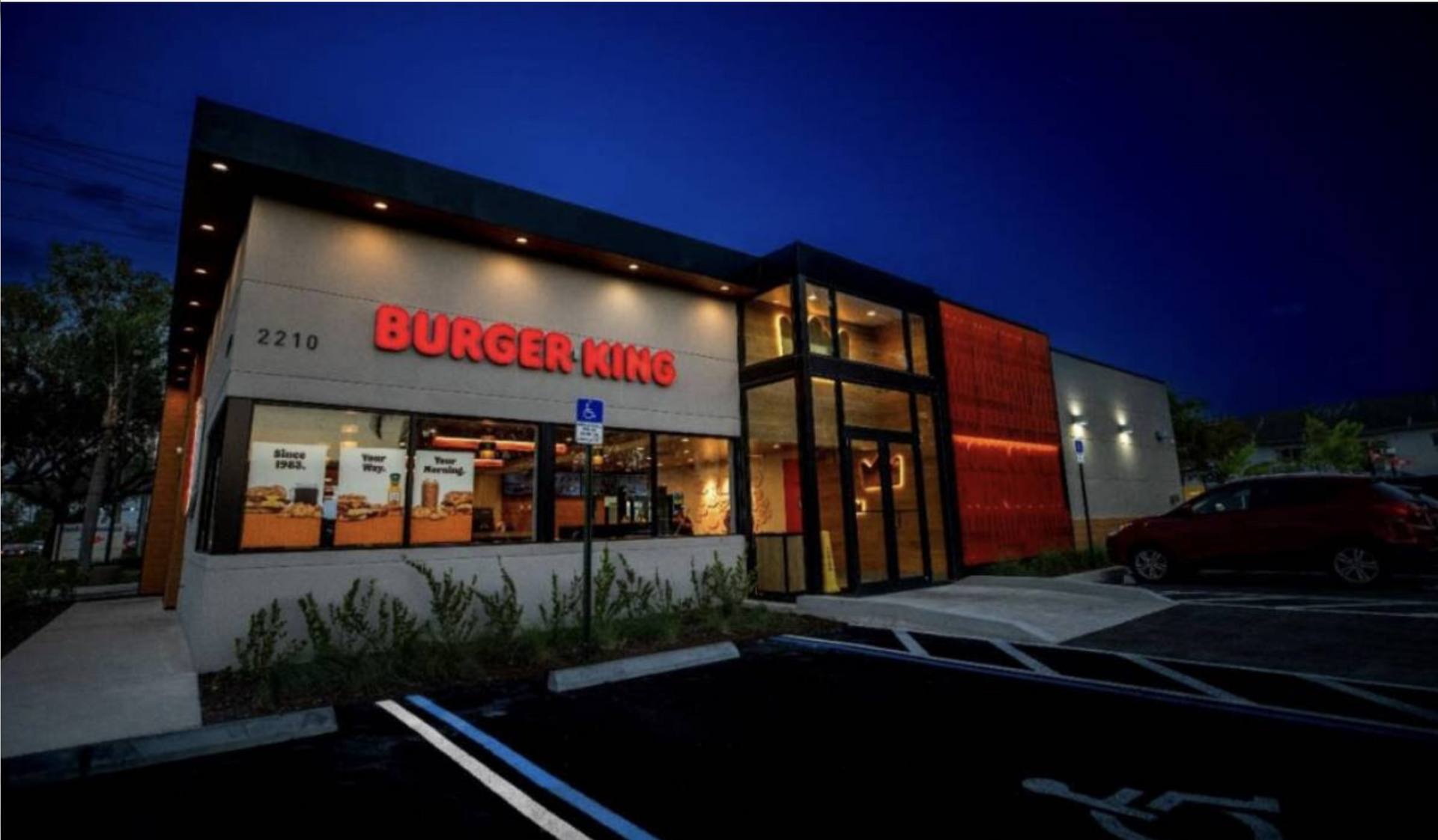 A burger king restaurant is lit up at night with cars parked in front of it.