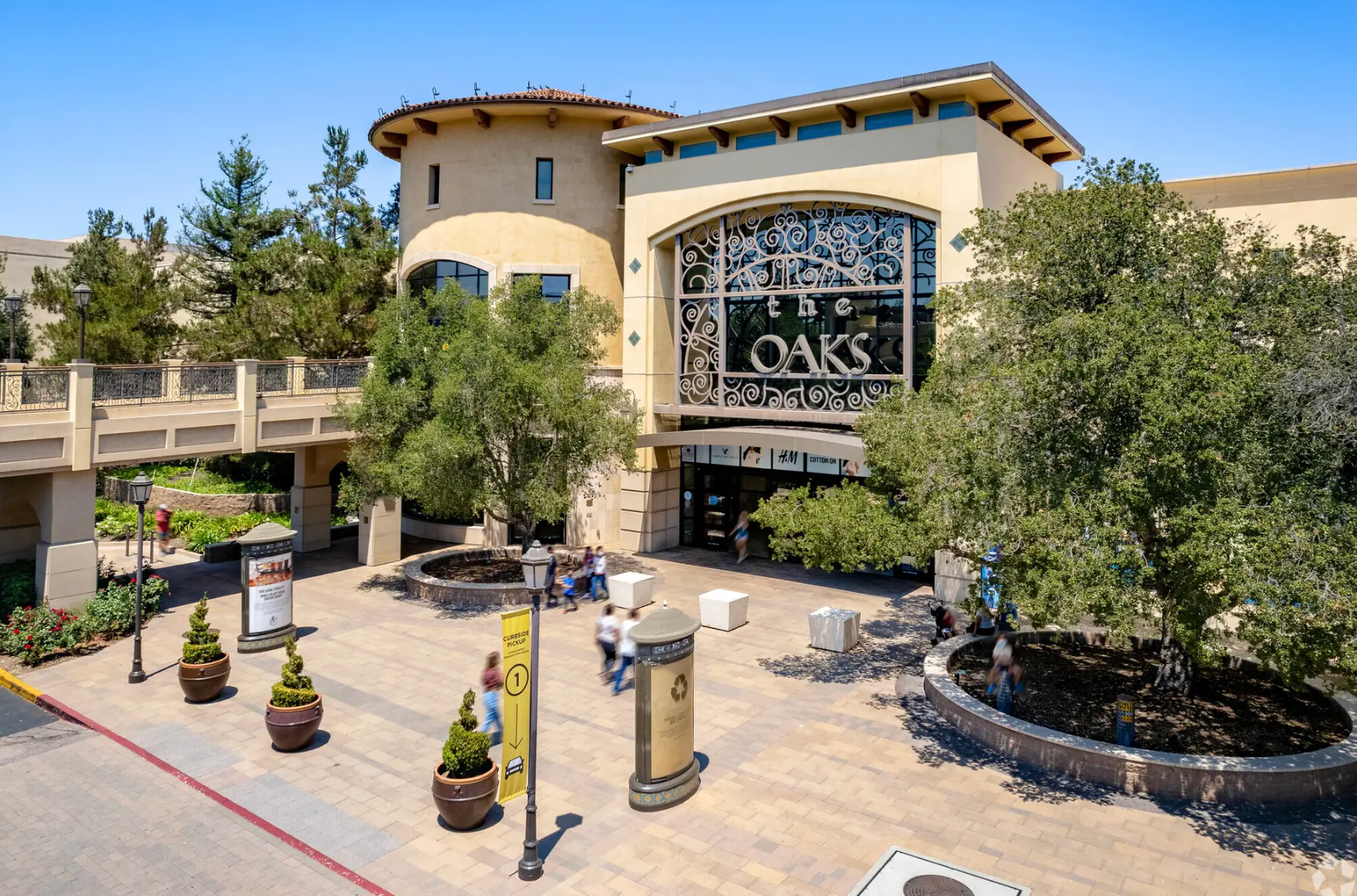 An elevated outdoor view of a modern shopping mall promenade with manicured greenery, palm trees, and pedestrians.