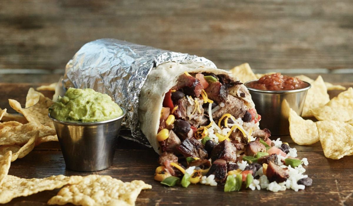A burrito is sitting on a wooden table next to tortilla chips and guacamole.