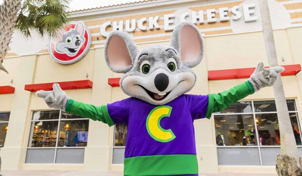 A chuck e cheese mascot is standing in front of a chuck e cheese restaurant.