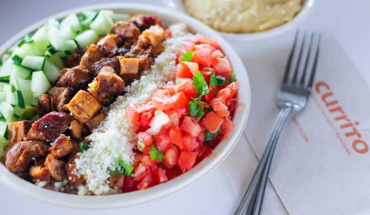 An elevated outdoor view of a modern shopping mall promenade with manicured greenery, palm trees, and pedestrians.