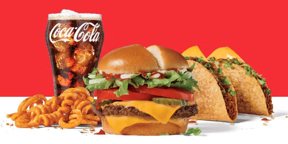 The American flag waves against a bright blue sky between towering glass skyscrapers, viewed from a low angle.