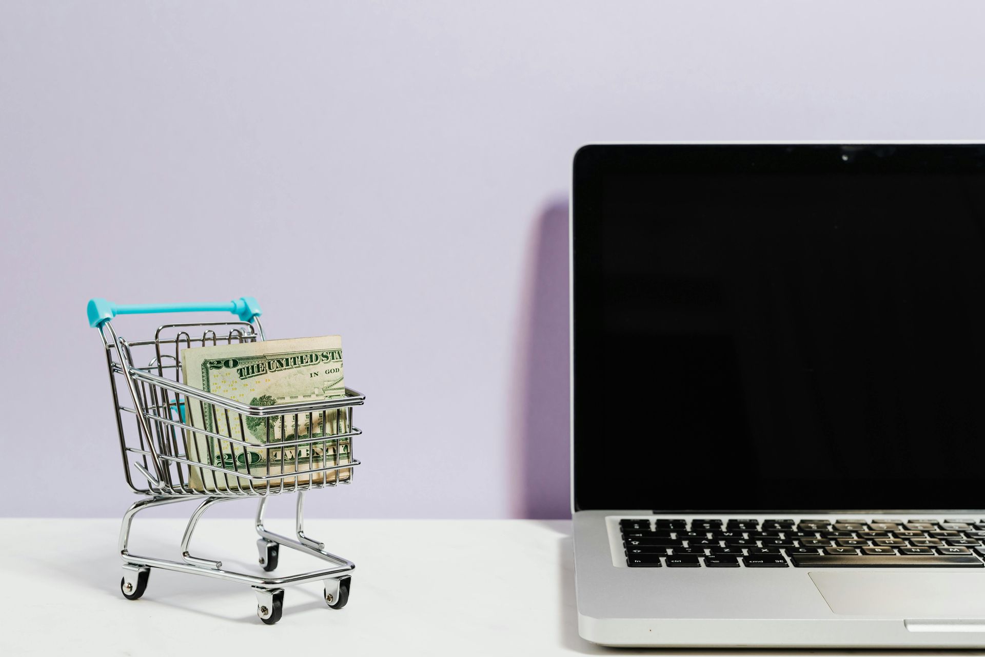 A shopping cart filled with money next to a laptop computer.
