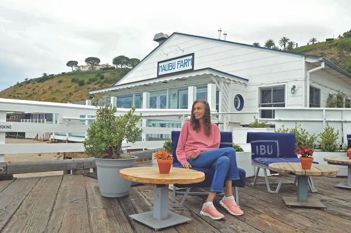 A woman is sitting at a table on a pier in front of a building.