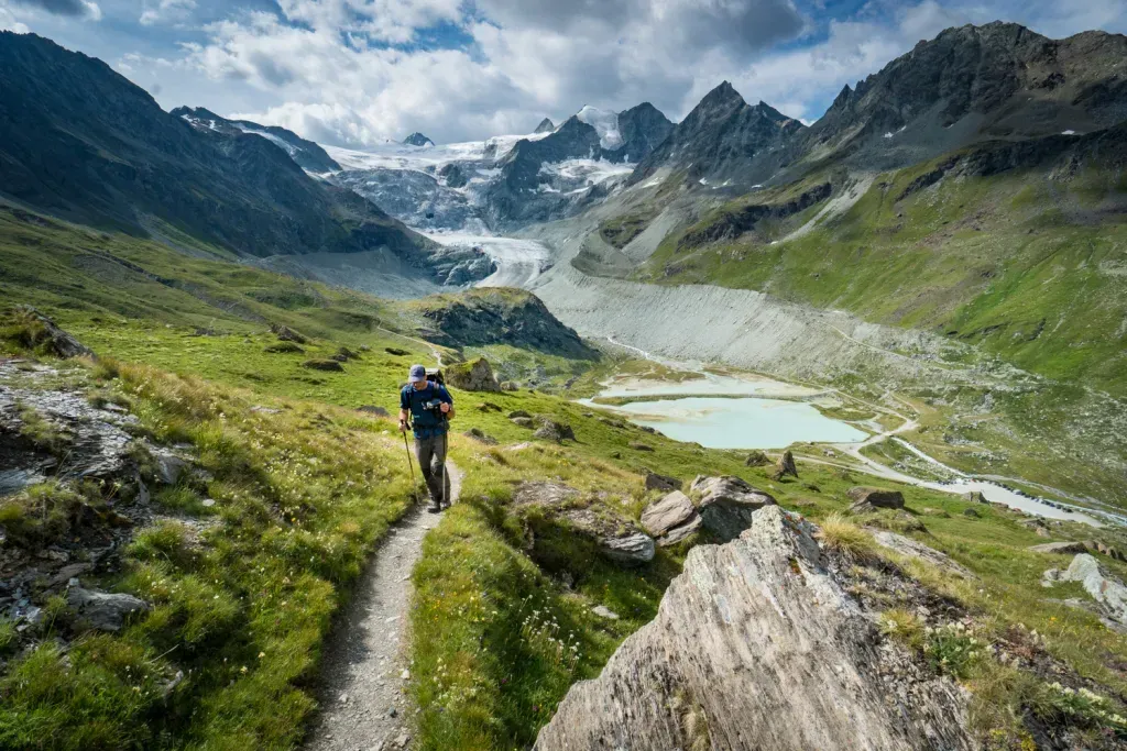 Hiker on a narrow mountain trail above a green valley and glacial lake under cloudy skies