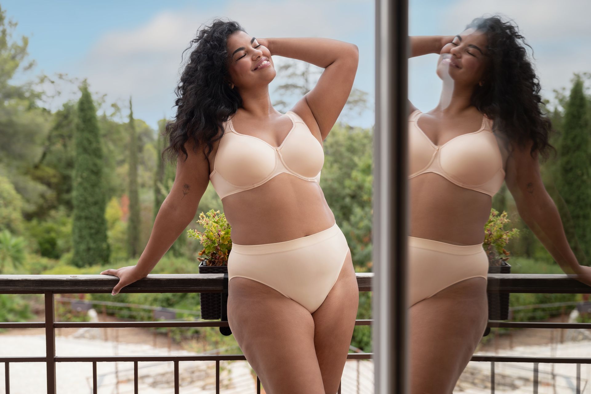 A woman in lingerie is standing on a balcony looking at her reflection in the mirror.