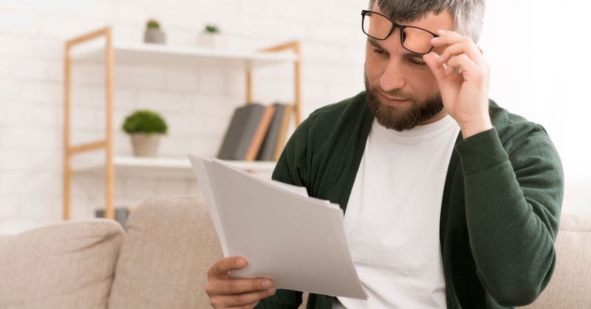 A middle-aged man on a couch lifts his glasses from his eyes to carefully examine the document he's holding.