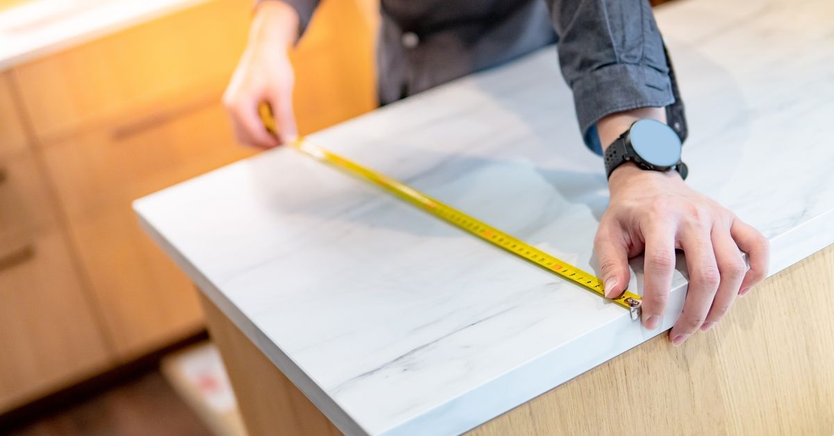 A close-up of a man stretching a tape measure across a marble kitchen countertop to establish its width.